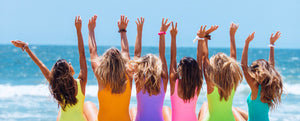 6 women with arms up at the beach facing the ocean