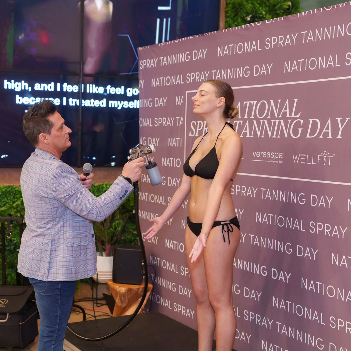 Woman in a black bikini receiving a spray tan from a man with a microphone at a National Spray Tanning Day event.