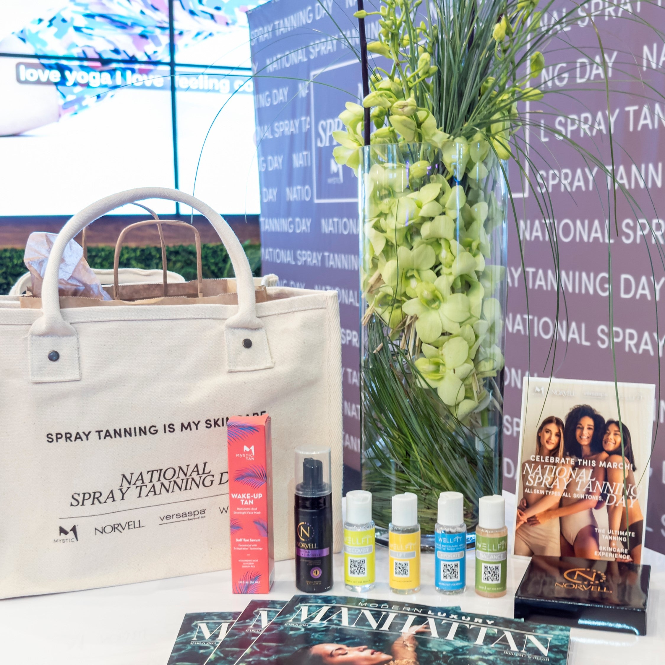 Spray tanning products and promotional materials on a table with a 'National Spray Tanning Day' banner in the background.